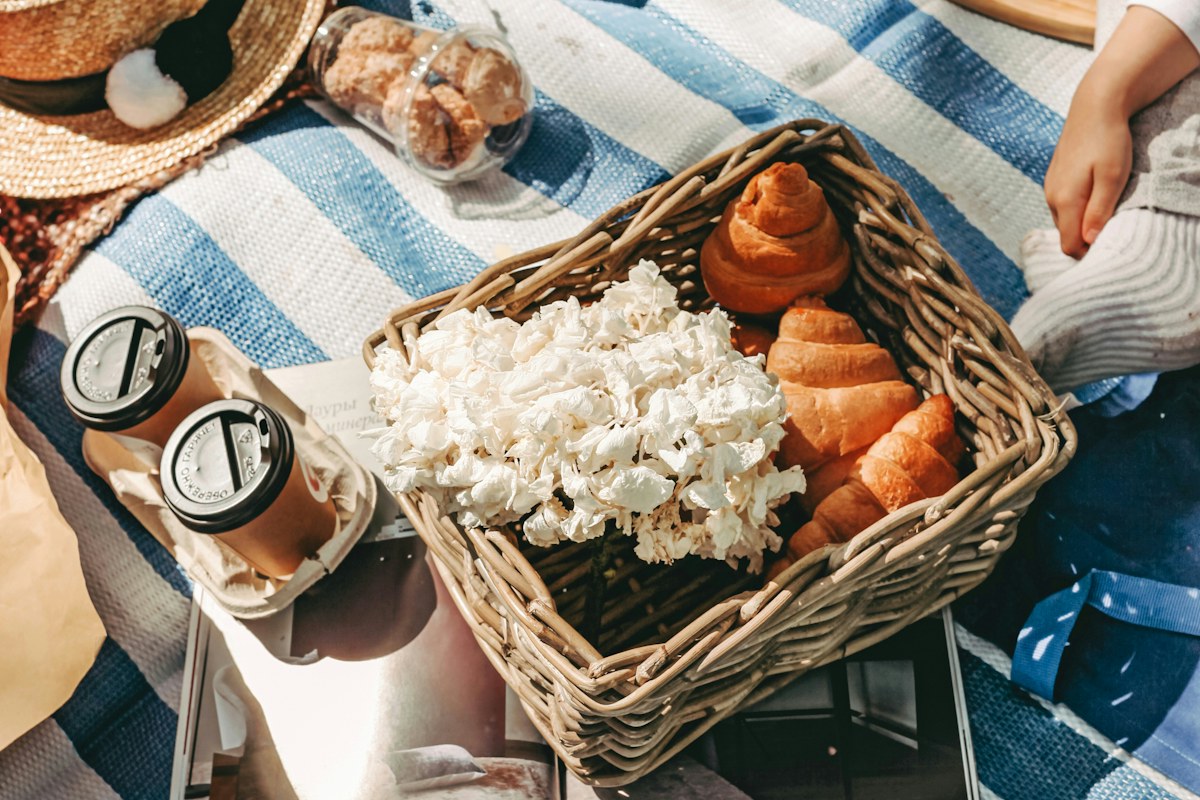 A wicker basket with picnic food and pale flowers on a striped blanket.