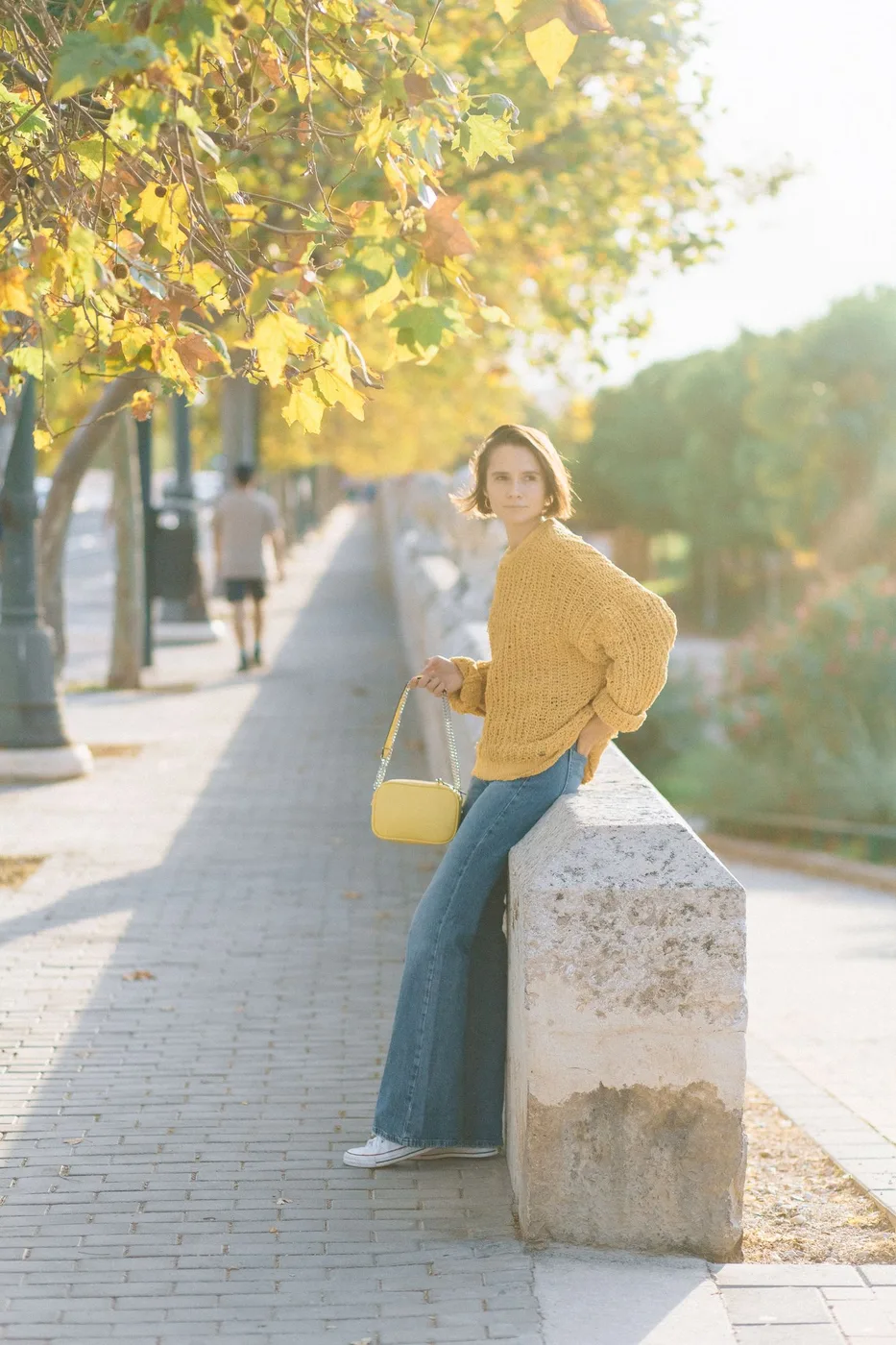 A woman in a yellow knit top and jeans beside a low outdoor wall.
