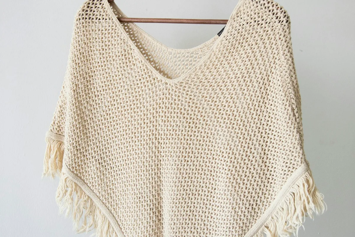 A pale open-textured knitted garment with fringed edges hanging against a light wall.
