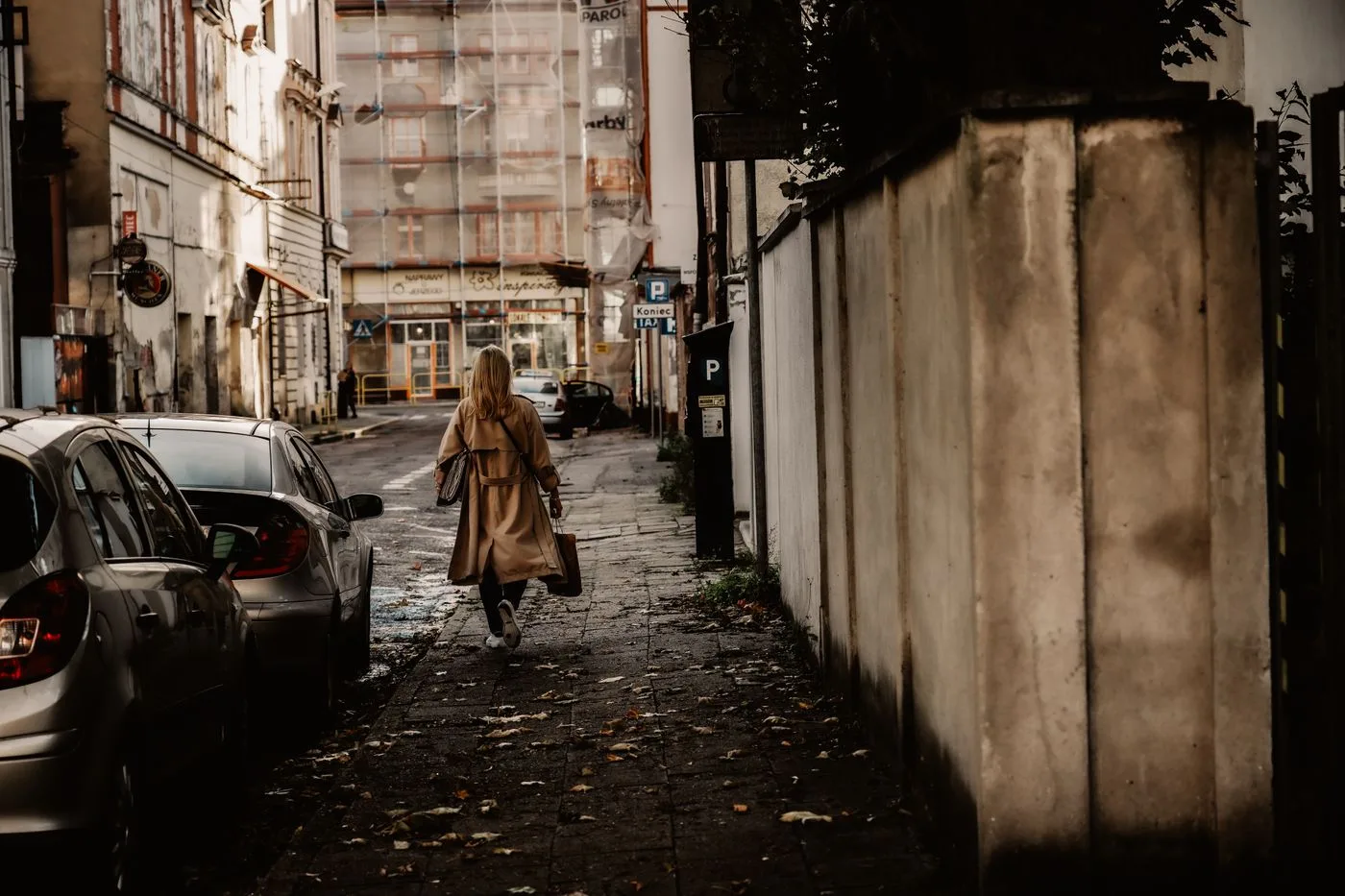 A person in a long brown outer layer walking away along a narrow city street.