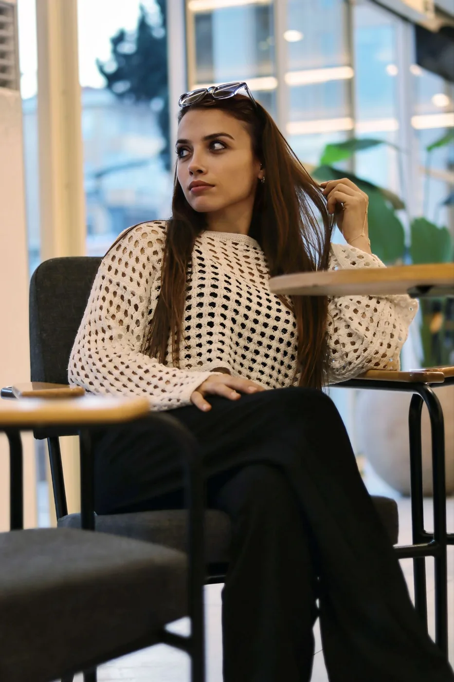 A woman in a pale open-textured knit over a dark layer with black trousers at a cafe table.