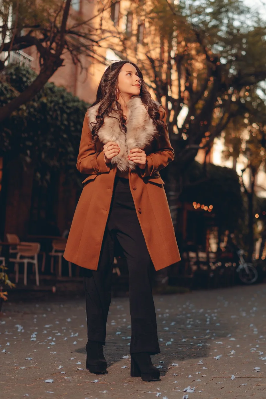 A woman wearing a brown coat and dark trousers among autumn trees.