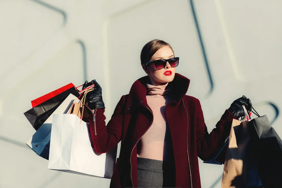 A woman in sunglasses and dark outerwear holding shopping bags.