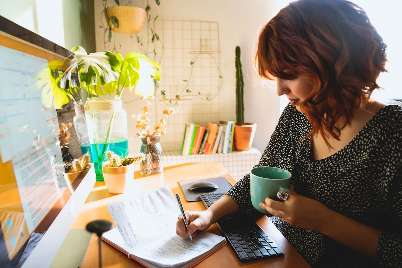 A woman writing in a notebook while holding a green mug at a desk.