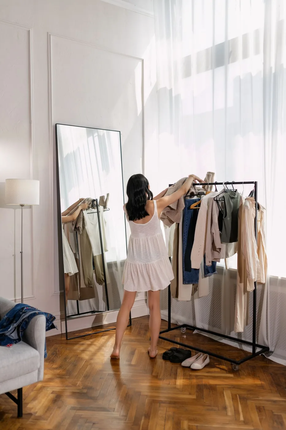A woman arranging clothes on a rail in a bright room with a floor mirror.