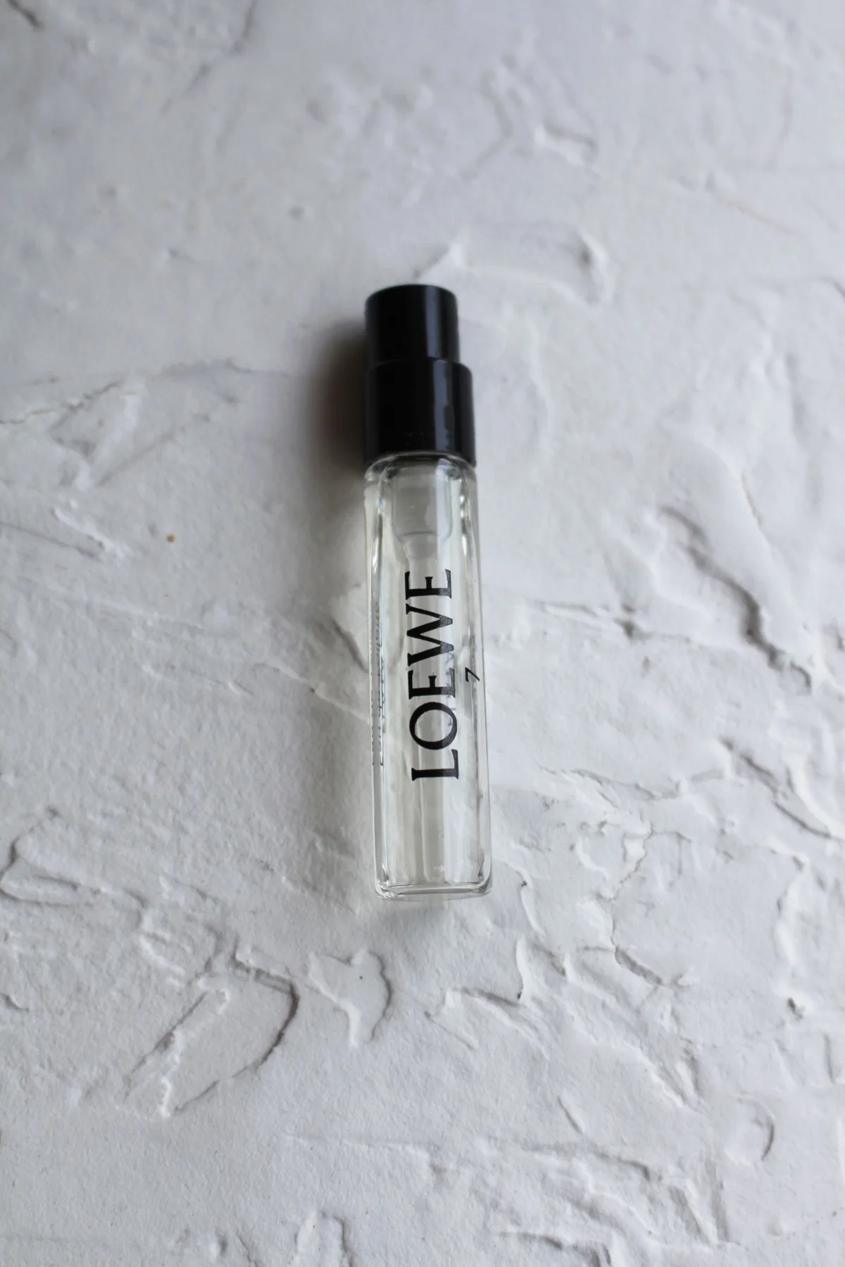 A small labelled perfume sample bottle on a pale textured surface.